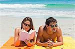 Tanned couple on the beach with the woman reading a book with the sea in background