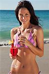 Smiling woman in a bikini stirs a cocktail using a straw while she stands on a beach
