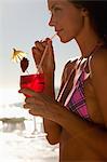 Woman wearing a bikini as she uses a straw to drink a cocktail while standing by the sea on a beach
