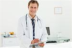 Smiling male doctor with clipboard and pen standing in his examination room