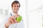 A smiling surgeon is holding a green apple while standing in front of a window