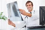 Arzt sitzt an seinem Schreibtisch eine Röntgenaufnahme und in die Kamera schaut lächelnd