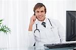 Smiling surgeon sitting at the desk behind his computer while using his mobile phone