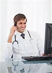 Doctor looking at his computer while sitting at the desk and using his cellphone