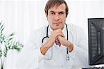 Thoughtful surgeon sitting behind his desk looking at the camera with his hands in front of his chin