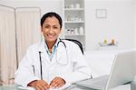 Smiling doctor sitting at the desk with a clipboard and a laptop in a medical office