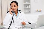 Serious doctor calling while sitting at her desk with a clipboard and a laptop