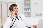 Relaxed doctor sitting at his desk while working on his laptop and calling