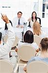 Man raising his hand as he sits in an audience while watching two business people who are sitting behind a table