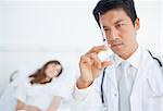 Serious doctor looking at a syringe as he prepares it while a patient is on a bed in the background