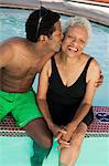 Man sitting on edge of swimming pool, kissing mother on cheek, elevated view.