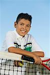 Young boy with tennis racket and ball leaning on tennis net, portrait
