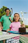 Father and Daughter by net on tennis court Holding Rackets and Trophy, portrait