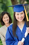 Graduate holding diploma with mother behind, outside, portrait