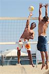 Young man jumping, hitting volleyball over net on beach; opponent defending