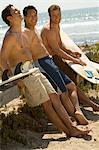 Three surfers leaning on bench holding surfboards on beach