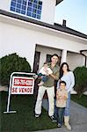 Family standing in front of house with For Sale sign, portrait