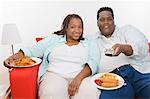 Mid-adult overweight couple sitting on sofa with meal and watching television