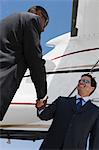 Two mid-adult businessman shaking hands in front of airplane, low angle view.