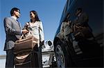 Mid-adult businesswoman and businessman standing in front of car with their luggage.