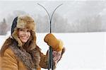Woman wearing fur coat and hat, holding skis, in snow covered field, portrait.