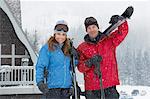 Couple holding skis, standing in front of cabin in snow