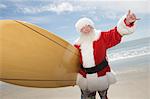 Père Noël se tient avec une planche de surf à la plage