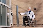 Doctor with muscular dystrophy in wheelchair at hospital entrance pressing knob for accessible door