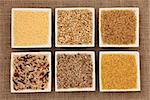 Cereal and grain selection of bulgur wheat, buckwheat, couscous, rye grain and brown and wild rice in white porcelain dishes on hessian sacking background.