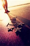 Three starfishes and word "India" on the sand. Woman's feet on the background