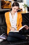 young girl student sitting on a sofa and reading a book,she watches a book with absorbed expression and takes pen with right hand