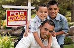 Hispanic Father and Sons in Front of a Sold Home For Sale Real Estate Sign.