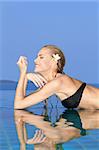 Smiling woman with wet hair laying with her arms on the tiled edge of the pool reflected in the water below