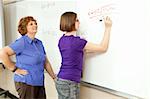 Student works algebra problem on the blackboard while her teacher looks on.