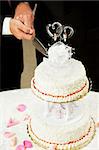 Closeup of a wedding cake topped with two grooms, being cut by two men's hands.