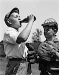 1930s PAIR OF BOYS WEARING BASEBALL GLOVES ONE WITH CATCHER'S MASK WATCHING OTHER WEARING CAP DRINK BOTTLE OF SODA