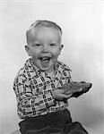 1950s LAUGHING BOY WEARING A PLAID SHIRT WITH JAM ON HIS FACE & MOUTH HOLDING A SLICE OF BREAD WITH BOTH HANDS LOOKING AT CAMERA
