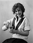 1920s SMILING YOUNG WOMAN SITTING POURING MILK FROM BOTTLE INTO CUP LOOKING AT CAMERA