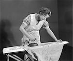 1930s TIRED WOMAN HOUSEWIFE WEARING APRON IRONING SHIRT WITH ELECTRIC IRON ON IRONING BOARD