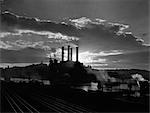 1930s SUNRISE SUNSET BEHIND SILHOUETTE OF INDUSTRIAL POWER PLANT WITH SMOKE STACKS NEAR RIVER