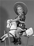 ANNÉES 1950 FILLE HABILLÉE EN COWGIRL CHEVAUCHANT LE CHEVAL À BASCULE