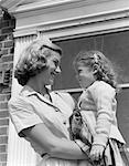 1950s MOTHER HOLDING HER DAUGHTER IN ARMS SMILING AT HER IN DOORWAY OUTSIDE OF THEIR HOME