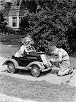 1940s 1930s BOY ON SIDEWALK FIXING HEADLIGHT OF TOY CAR DRIVEN BY LITTLE GIRL PLAYING OUTDOOR