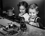 DES ANNÉES 1950 GARÇON & FILLE DEBOUT AU TABLEAU REGARDER TOY TRAIN VA SUR LA PISTE DE