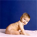 1980s NAKED BABY SITTING ON PINK BLANKET AGAINST DARK BLUE BACKGROUND PROFILE LEANING FORWARD LOOKING AT CAMERA OVER SHOULDER
