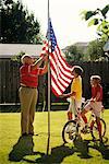 GRANDFATHER RAISING FLAG ON POLE TWO GRANDCHILDREN WATCHING