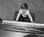 1980s OVERHEAD OF BOY SITTING ON BENCH WITH FINGERS ON PIANO KEYS LOOKING UP