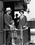 ANNÉES 1930 FAMILLE DE TROIS PERSONNES DEBOUT AU GARDE-CORPS À L'ARRIÈRE DE LA VOITURE DE TRAIN AVEC PORTEURS EN ARRIÈRE-PLAN