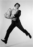 1960s MAN IN SUIT & FEDORA LEAPING FORWARD WITH GROCERY BAGS IN ARMS LOOKING AT CAMERA