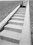 1960s BUSINESSMAN CROUCHED IN MIDDLE OF LONG FLIGHT OF CEMENT STAIRS WITH BRIEFCASE SET DOWN IN FRONT OF HIM